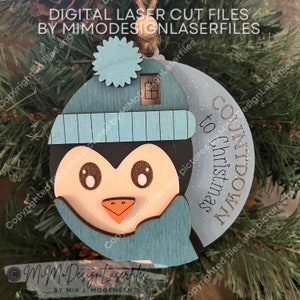Può includere: Un ornamento di pinguino in legno blu e bianco con un cappello e una sciarpa blu. L'ornamento ha il testo "COUNTDOWN TO CHRISTMAS".
