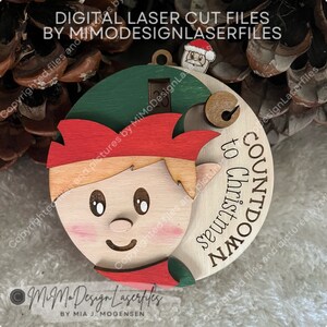 Può includere: Un ornamento di Natale in legno a forma di conto alla rovescia con un elfo con un cappello rosso, una camicia verde e una barba marrone. L'ornamento ha un ritaglio per un numero e il testo "COUNTDOWN TO CHRISTMAS" è scritto sul lato.