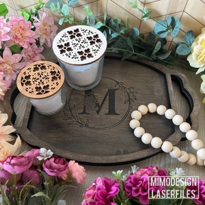 Può includere: Un vassoio di legno con un bordo nero e un motivo floreale inciso al centro. Il vassoio ha due manici e presenta una grande lettera "M" al centro. Ci sono due candele con coperchi decorativi sul vassoio e una collana di perline di legno.