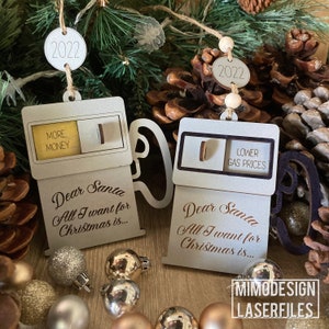 May include: Two wooden Christmas ornaments shaped like gas pumps. The ornaments are white with brown accents. The top of each ornament has a circle with the year 2022 written on it. The left ornament has a yellow rectangle with the words "More Money" written on it. The right ornament has a black rectangle with the words "Lower Gas Prices" written on it. Both ornaments have the text "Dear Santa All I want for Christmas is..." written on them.