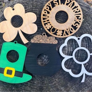 May include: Five wooden ornaments for St. Patrick's Day. The ornaments are shaped like a shamrock, a pot of gold, a leprechaun hat, a shamrock flower, and a circle with the text "Happy St. Patrick's Day".