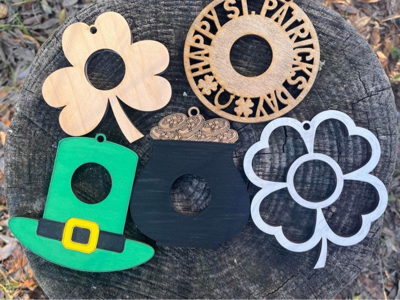 May include: Five wooden ornaments for St. Patrick's Day. The ornaments are shaped like a shamrock, a pot of gold, a leprechaun hat, a shamrock flower, and a circle with the text "Happy St. Patrick's Day".