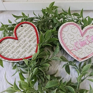 May include: Two heart-shaped Valentine's Day ornaments. One has a red border and the words "Happy Valentine's Day" printed on it. The other has a pink border and the words "I love you" in cursive. Both have a small hole for hanging.