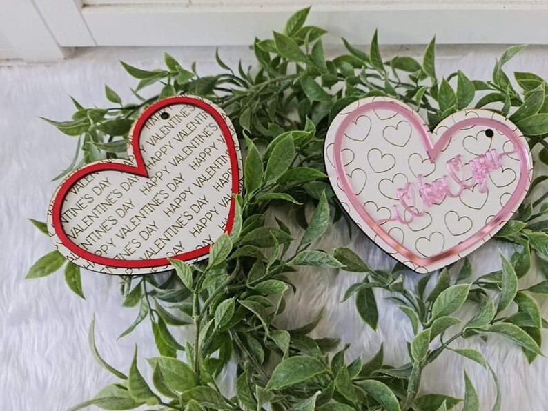 May include: Two heart-shaped Valentine's Day ornaments. One has a red border and the words "Happy Valentine's Day" printed on it. The other has a pink border and the words "I love you" in cursive. Both have a small hole for hanging.