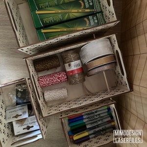 May include: Wooden storage boxes with decorative cutouts, filled with craft supplies. The boxes contain pencils, twine, ribbon, and markers. The boxes are stacked on top of each other.