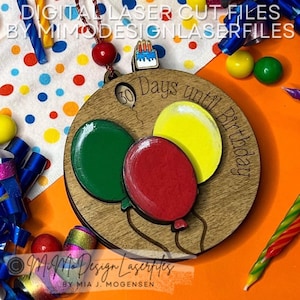 Puede incluir: Un adorno redondo de madera con una cuenta atrás para el cumpleaños. El adorno presenta tres globos en colores rojo, verde y amarillo. El texto en el adorno dice "30 días hasta el cumpleaños".