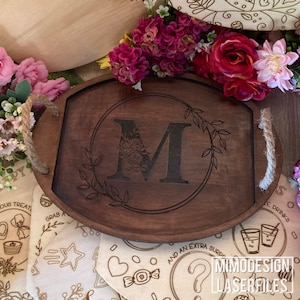 Puede incluir: Una bandeja de servir de madera con un asa de cuerda, que presenta un diseño grabado con láser de una corona floral con la letra "M" en el centro. La bandeja es marrón y tiene un acabado de madera natural.