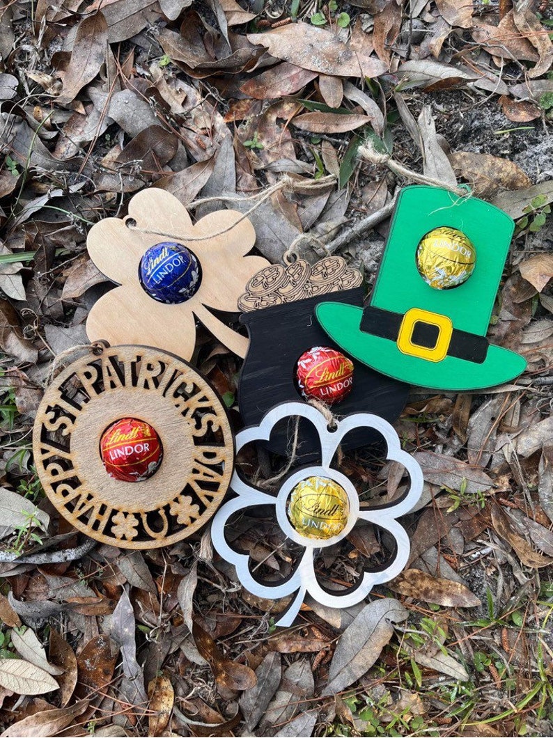 May include: Five wooden ornaments with a shamrock, pot of gold, leprechaun hat, and two other shamrocks. Each ornament has a small chocolate candy in the center.
