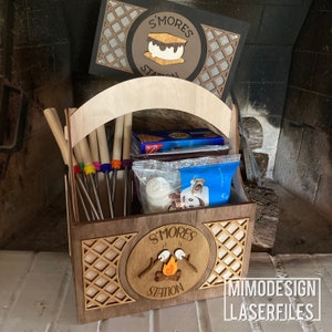 Può includere: Stazione di marshmallow in legno con manico, con un cerchio marrone con il testo "S'mores Station" e una grafica di un falò. La stazione include un set di spiedini in metallo, una confezione di cracker graham, una confezione di barrette di cioccolato e un sacchetto di marshmallow.