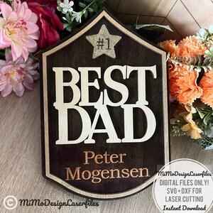 May include: A dark brown wooden plaque with a gold star and the text "#1 BEST DAD Peter Mogensen" in white and gold lettering. The plaque has a decorative border and is suitable for laser cutting.