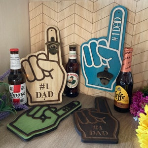May include: Wooden and painted foam finger bottle openers with the text "#1 DAD" and "OPEN HERE". The openers are in natural wood, blue, green, and brown. Several beer bottles are also visible.