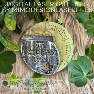 Può includere: Un ornamento rotondo in legno con un design a forma di pentola d'oro. L'ornamento è dipinto in tonalità di verde, oro e nero. Il testo "Counting down to St. Patrick's Day" è scritto sull'ornamento. Il numero "14" è visibile in un ritaglio sull'ornamento.