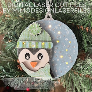 May include: A light blue wooden ornament with a penguin wearing a green and blue hat. The ornament has a white and silver glitter design and is decorated with small white dots. The ornament is hanging on a Christmas tree.