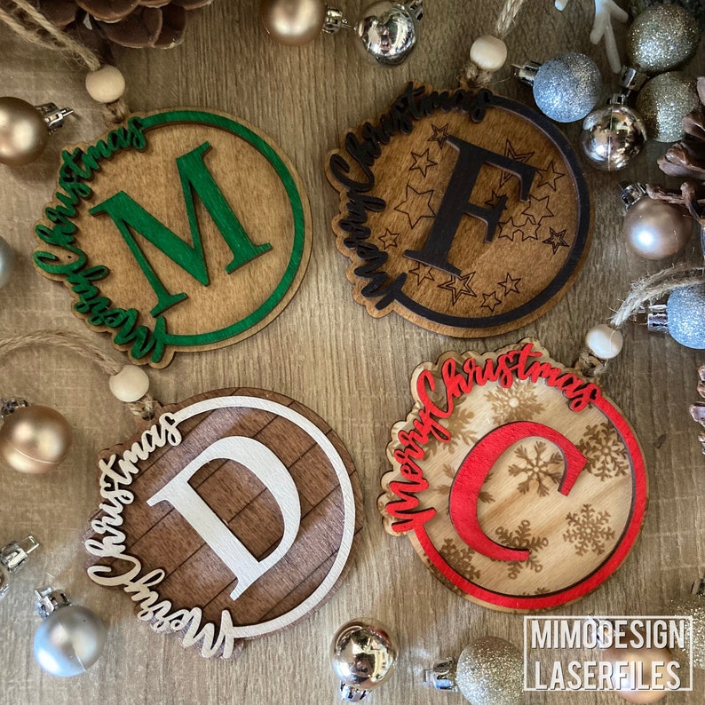 Op de afbeelding: Vier houten kerstversieringen met verschillende letters en ontwerpen. De versieringen zijn rond en hebben een rustieke houtnerfstructuur. De letters zijn M, F, D en C. De ontwerpen omvatten sneeuwvlokken, sterren en een houtnerfpatroon.