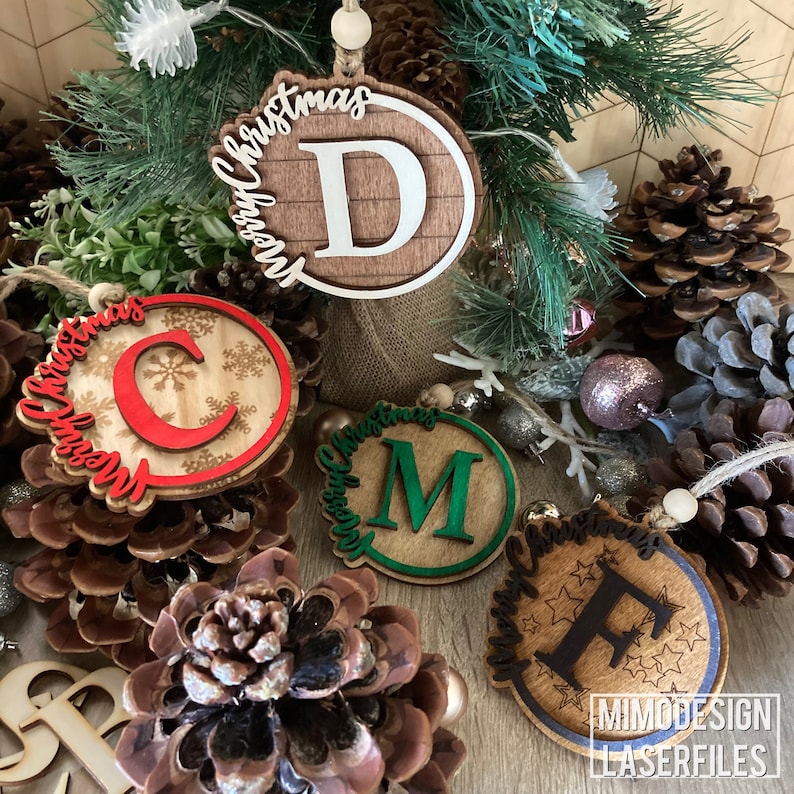 Op de afbeelding: Vijf houten kerstversieringen met de letters C, D, M en F in het midden van elke versiering. De versieringen zijn versierd met sneeuwvlokken, sterren en de woorden "Merry Christmas".