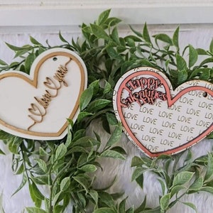 May include: Two heart-shaped wooden ornaments. One has "I love you" in cursive, the other says "Happy Valentine's Day" with a "love" pattern. Both are surrounded by a gold border and set against a backdrop of greenery.