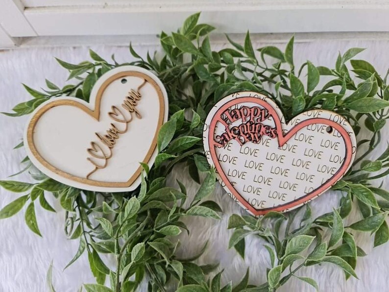 May include: Two heart-shaped wooden ornaments. One has "I love you" in cursive, the other says "Happy Valentine's Day" with a "love" pattern. Both are surrounded by a gold border and set against a backdrop of greenery.