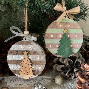 May include: Two wooden Christmas baubles with a light-up tree design. One bauble is brown with silver glitter and the other is green with pink stripes. Both baubles have a gold star on top of the tree.