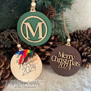 Può includere: Tre ornamenti rotondi in legno con diversi design. Il primo ornamento è verde con una lettera dorata "M" al centro. Il secondo ornamento è in legno naturale con schizzi di vernice colorata e il testo "Feliz 2024". Il terzo ornamento è marrone con il testo "Merry Christmas 2024".
