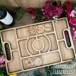 Op de afbeelding: Een houten serveerblad met een uitgesneden ontwerp en meerdere compartimenten. Het blad heeft een handvat aan de zijkant en heeft verschillende ronde houten schijven met gegraveerde tekst, waaronder "Wine Down", "Tapas", "Wine & Dine" en "Shark Coochie Board".