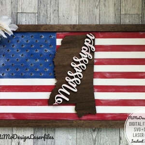 May include: A wooden sign with a red, white, and blue American flag design. The state of Mississippi is cut out of the flag and the word "Mississippi" is written in pink cursive letters on the state outline. The sign is framed in a dark brown wood frame.