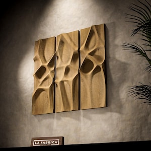 May include: A triptych of three vertical light brown wooden panels featuring deep, organic, wave-like carved indentations. A small rectangular sign with the text LA FABRICA is positioned below the panels on a dark surface.