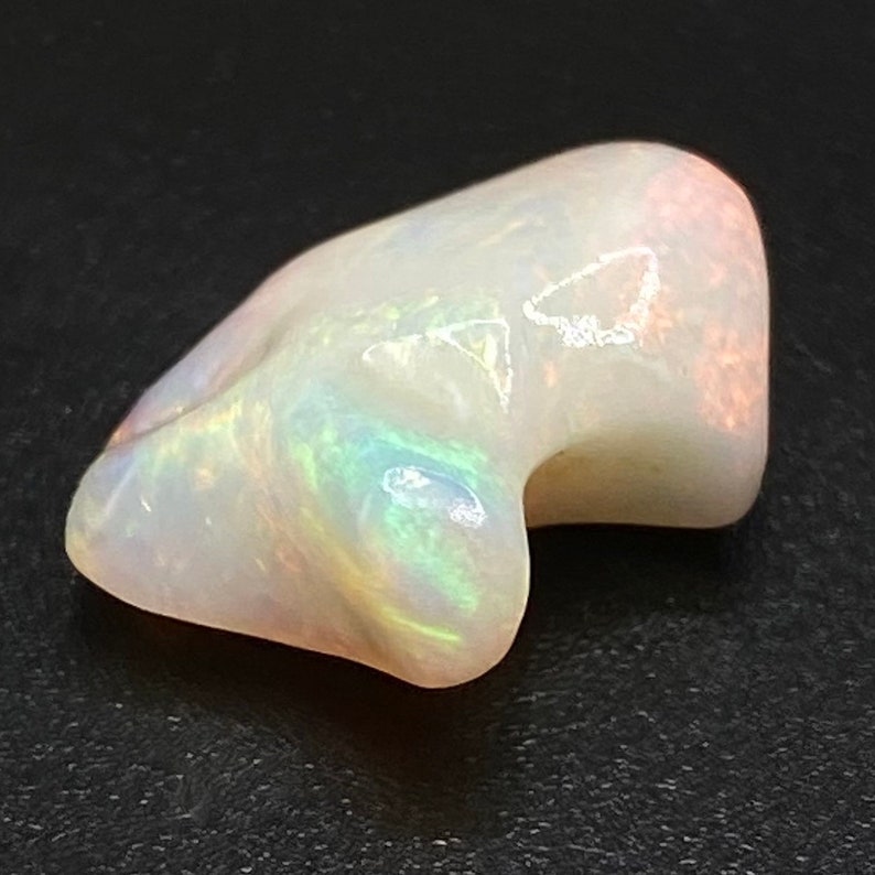 May include: A white opal gemstone with iridescent green, blue, and pink flashes. The opal is irregularly shaped and has a smooth, polished surface.