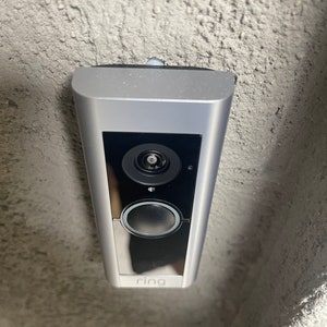 May include: A silver Ring doorbell mounted on a textured gray wall. The doorbell has a black camera lens and a black button.