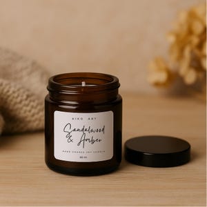 Op de afbeelding: Een bruine glazen potkaars met een zwarte deksel. Het etiket luidt "Sandalwood & Amber" en "Hand Poured Soy Candle". De kaars is 60 ml. De pot staat op een houten oppervlak.
