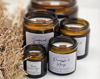 May include: Four brown glass jar candles with white labels and wooden wicks. The labels read "Sandalwood & Amber", "Pineapple & Mango", and "Baked Cinnamon Bun". The candles are arranged in a group, showcasing different sizes.