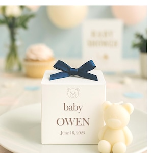 May include: A white square gift box topped with a dark blue satin ribbon bow. The front of the box features a line art bear icon above the printed text baby OWEN and the date 18 June 2025. A small cream-coloured bear-shaped candle sits beside it.