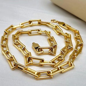 Könnte beinhalten: Eine goldfarbene Kettenhalskette mit einer Reihe von länglichen, rechteckigen Gliedern, die in einem sich wiederholenden Muster verbunden sind. Die Kette verfügt über einen Karabinerverschluss und eine polierte, reflektierende Oberfläche.