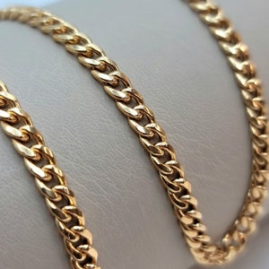 May include: A gold-colored curb chain necklace. The chain is made of interlocking links and is displayed on a neutral-toned surface. The chain has a polished finish, reflecting light.