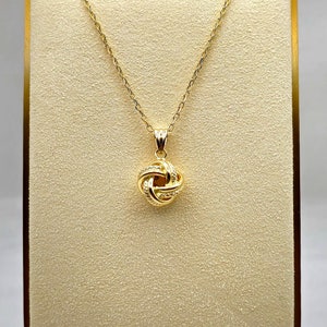 May include: A gold-colored necklace with a pendant in the shape of a woven knot. The pendant is suspended from a delicate chain. The necklace is displayed on a beige stand with a gold-colored frame.