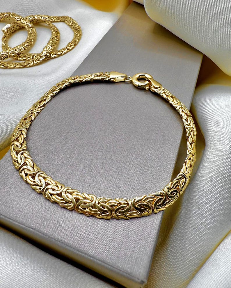 May include: A gold bracelet with a woven pattern, displayed on a grey surface. The bracelet is circular, with a clasp fastening. A matching gold necklace is visible in the background. Jewellery.
