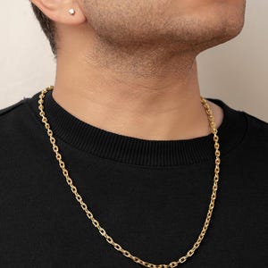 Puede incluir: Un collar de cadena de oro con eslabones ovalados de acabado pulido, usado alrededor del cuello sobre una camiseta negra de cuello redondo. Un peque&ntilde;o pendiente de bot&oacute;n redondo y transparente es visible en el l&oacute;bulo.