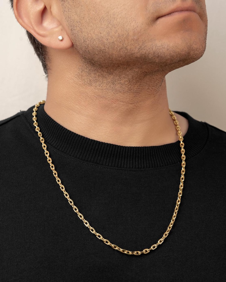 Puede incluir: Un collar de cadena de oro con eslabones ovalados de acabado pulido, usado alrededor del cuello sobre una camiseta negra de cuello redondo. Un peque&ntilde;o pendiente de bot&oacute;n redondo y transparente es visible en el l&oacute;bulo.