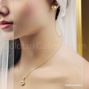 May include: A gold-colored jewelry set featuring a necklace and matching earrings. The necklace has a delicate chain with a knot-shaped pendant. The earrings are also knot-shaped. The jewelry is displayed on a mannequin.