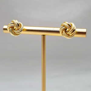 May include: A pair of gold-colored knot earrings displayed on a gold stand. The earrings have a textured, woven design. The background is a soft, neutral gray.