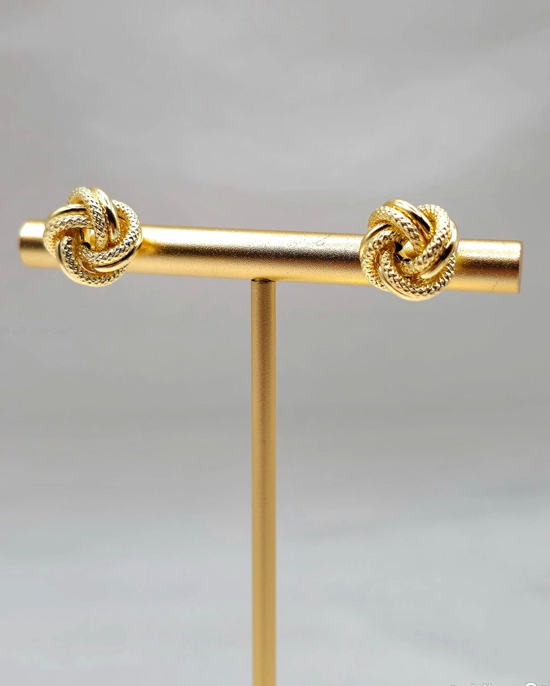 May include: A pair of gold-colored knot earrings displayed on a gold stand. The earrings have a textured, woven design. The background is a soft, neutral gray.