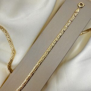 May include: A gold bracelet featuring a Byzantine chain pattern. The bracelet is presented on a neutral rectangular surface, with a cream-colored fabric background. The bracelet has a clasp for secure fastening.