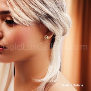 May include: Close-up of a person wearing a pair of gold knot earrings. The earrings are small, detailed, and appear to be made of gold. The background is blurred, with the text "GoldLux Galleria.com" visible.