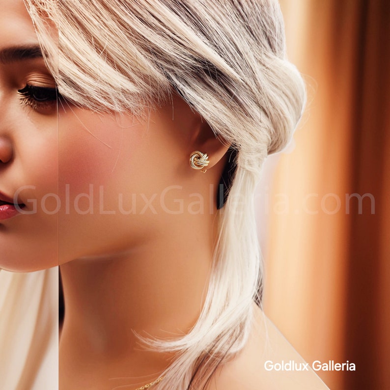 May include: Close-up of a person wearing a pair of gold knot earrings. The earrings are small, detailed, and appear to be made of gold. The background is blurred, with the text "GoldLux Galleria.com" visible.
