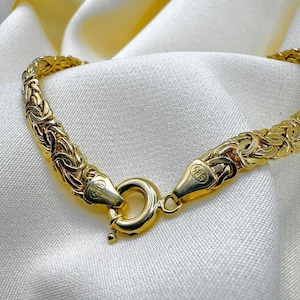 May include: A gold-coloured bracelet with a woven design, shown close-up. The bracelet is fastened with a round clasp. The background is a white, silky fabric.