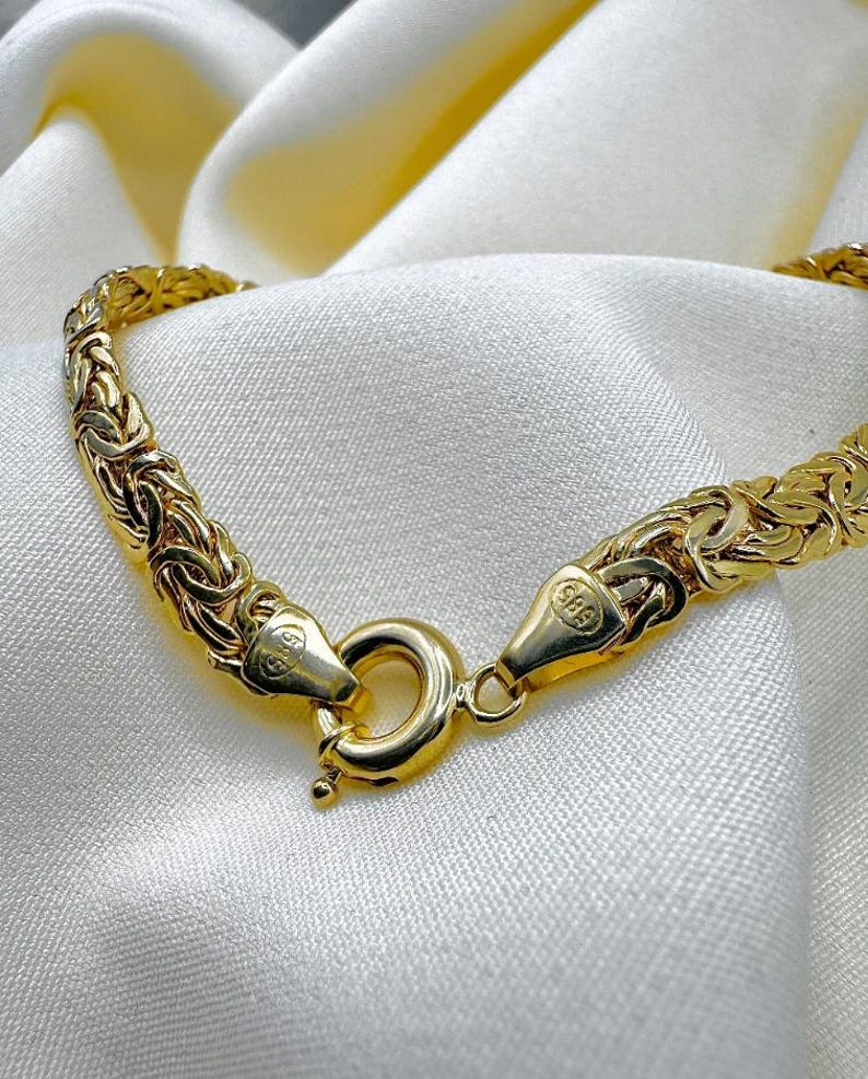 May include: A gold-coloured bracelet with a woven design, shown close-up. The bracelet is fastened with a round clasp. The background is a white, silky fabric.