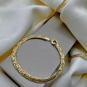 May include: A gold bracelet and necklace set with a woven design. The jewellery is displayed on a grey box and white satin fabric. The bracelet has a clasp fastening. The necklace is partially visible.