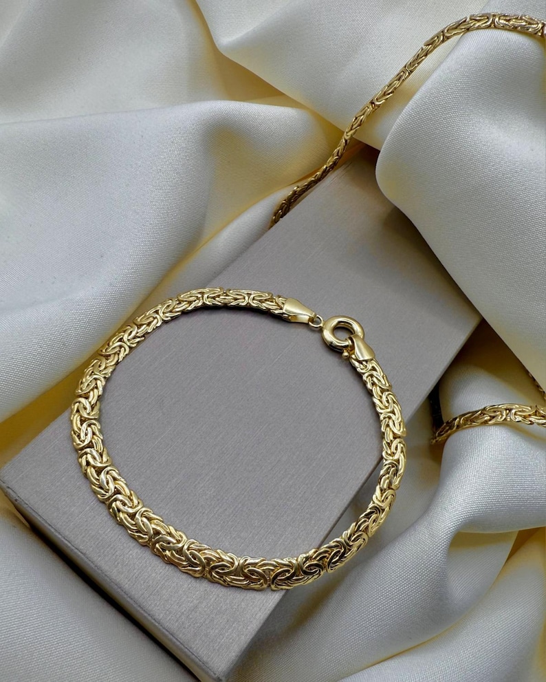 May include: A gold bracelet and necklace set with a woven design. The jewellery is displayed on a grey box and white satin fabric. The bracelet has a clasp fastening. The necklace is partially visible.