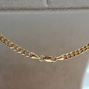 May include: Close-up of a gold curb chain necklace. The necklace features a series of interlocking links and a secure clasp. The metal has a bright, polished finish, reflecting light. The clasp is marked with "750".