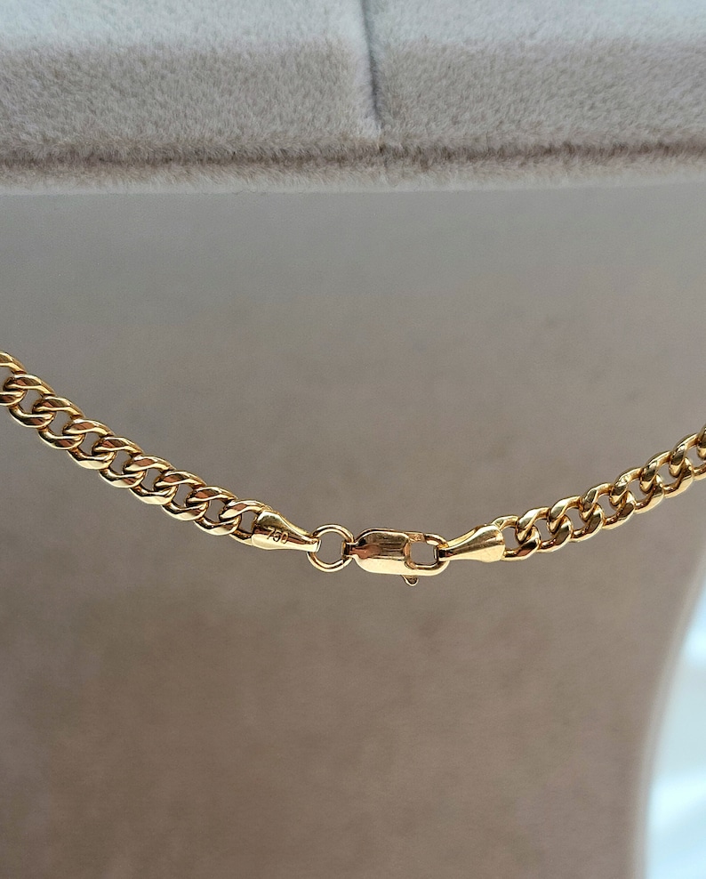 May include: Close-up of a gold curb chain necklace. The necklace features a series of interlocking links and a secure clasp. The metal has a bright, polished finish, reflecting light. The clasp is marked with "750".