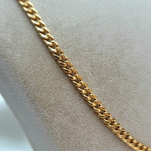 May include: A gold-colored curb chain necklace. The chain is made of interlocking links, creating a textured surface. The necklace is displayed against a neutral background, highlighting its shiny, metallic appearance. The chain is a classic jewelry piece.